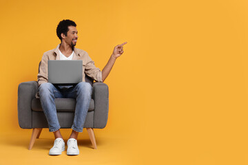 Happy man with laptop sitting in armchair on orange background. Space for text
