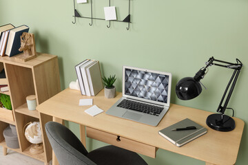 Modern laptop, books, lamp and stationery on wooden desk near green wall. Home office