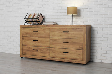 Wooden chest of drawers, books and lamp near white brick wall. Interior design