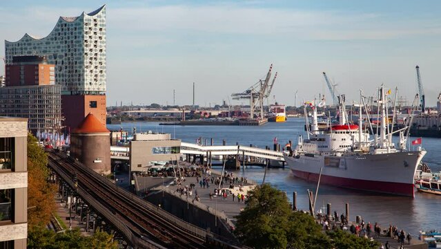 Hamburg cityscape timelapse featuring elevated railway, landmark skyline, Elbe river, a large ship at the pier, and a bustling promenade with crowds during a clear autumn day.