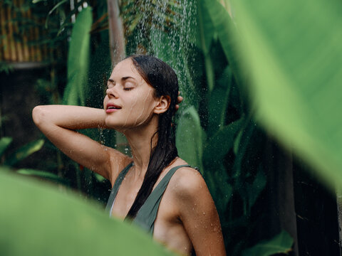 A woman in a swimsuit washes her head in a tropical shower outdoors against the backdrop of green tropical leaves, flowers and palm trees. Body and hair care, tanned skin, sexy, open mouth