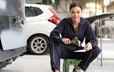 Woman auto mechanic putty damaged car body. Female repair technician prepare for painting crash...