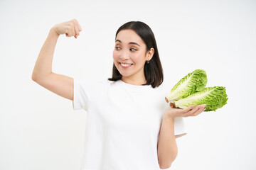 Strong asian woman shows fresh vegetables, lettuce and her muscles, flexing biceps with smiling...