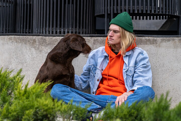 Upset depressed millennial guy sociopath sitting on ground outdoors with his dog. Unhappy lonely...