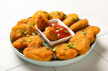 Plate with delicious nuggets, bowl of ketchup and cut green onion on white tiled table