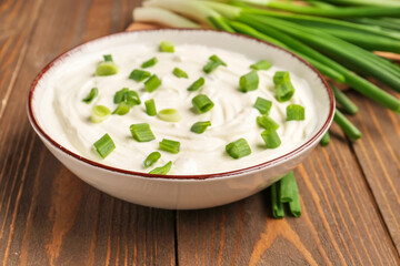 Bowl of tasty sour cream with green onion on wooden background, closeup