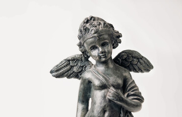Cherub or little angle figurine on light grey background. Partial body of metal boy with wings looking at camera with serene or peaceful expression. Spirituality or love background. Selective focus.