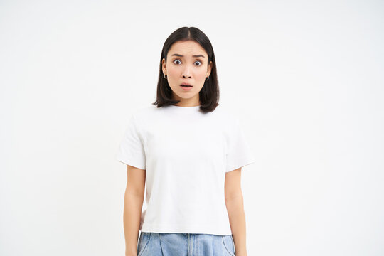 Image of concerned japanese woman, gasps and stares with shock at camera, poses in white t-shirt against studio background