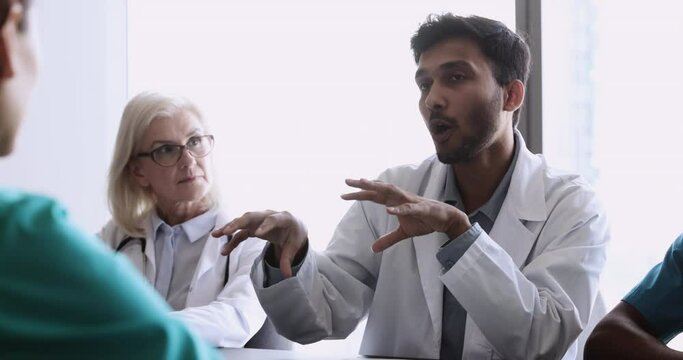 Young 35s Indian doctor talking to colleagues gathered in boardroom during medical council event or conference in clinic, to discuss diagnosis, prognosis, share opinion about patient further treatment