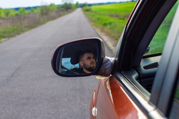 A man carefully look ahead while driving a car and is reflected in the mirror. Against the backdrop of a beautiful rural landscape, a man drives a car. The man's face is reflected in the mirror.