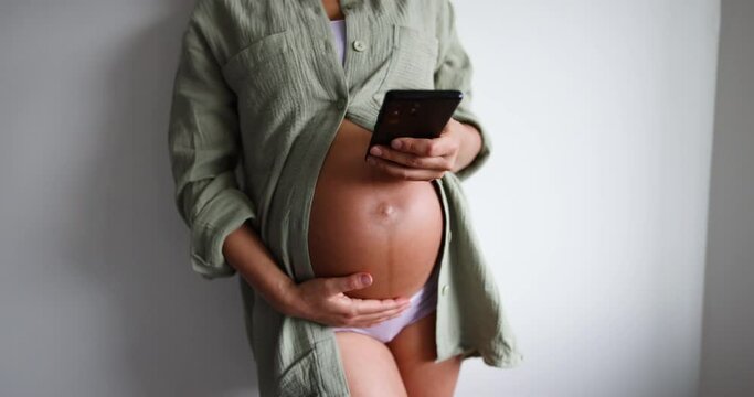 Pregnant woman using phone app for online pregnancy prenatal class or reading about motherhood wearing underwear and casual cotton shirt at home. Relaxing holding expecting belly for health