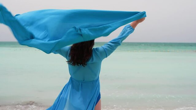 Woman in turquoise airy blue dress tunic with pareo over head stands on ocean beach in windy day admires waves, back view. Brunette female enjoying summer holidays on tropical resort. Travel, tourism.