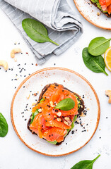 Avocado and salmon toast on rye bread with spinach leaves, cashew nuts and sesame seeds, white table background, top view