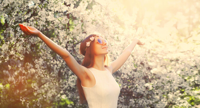 Beautiful young hippie woman raising her hands up wearing floral headband, sunglasses in spring blooming garden on white flowers tree background - Powered by Adobe