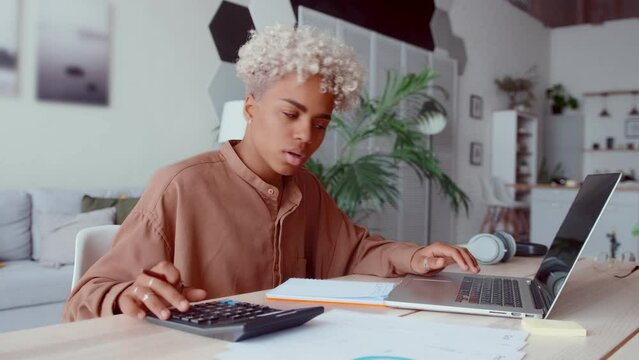 Young smart African American woman accountant uses calculator and makes notes in notebook and laptop compiling financial report and preparing for exam in economic college sits at table in home office.