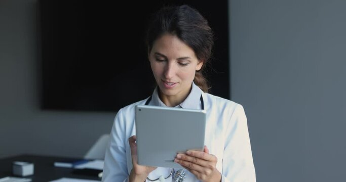 Smiling attractive Latina female doctor in white medical uniform use tablet gadget in modern private hospital, looking at pad screen, consult client on-line, fill patient medical history or anamnesis