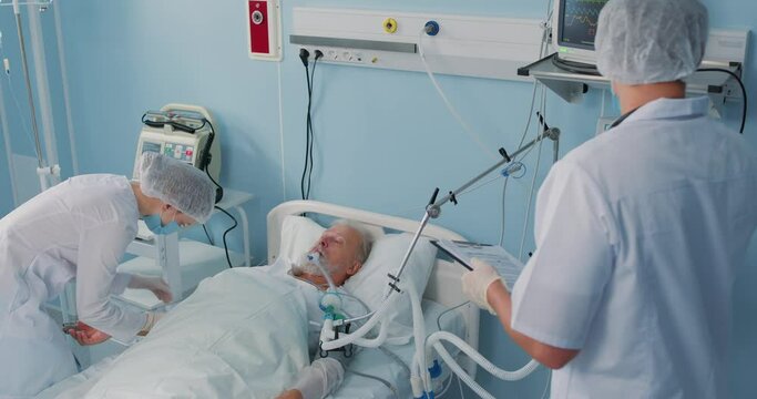 Sick old senior man patient resting in bed while assistants analyzing rehabilitation expertise, slow motion. Emergency in Hospital. Doctor Holds Hand to Check Heart Rate. Doctors visiting old sick man