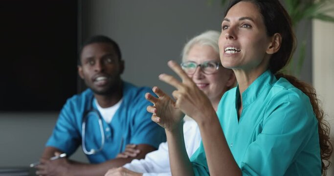 Different age and race physicians take part in meeting at clinic boardroom. Medical workers in uniform listening to Latina female general practitioner talks to workmates during medical council event