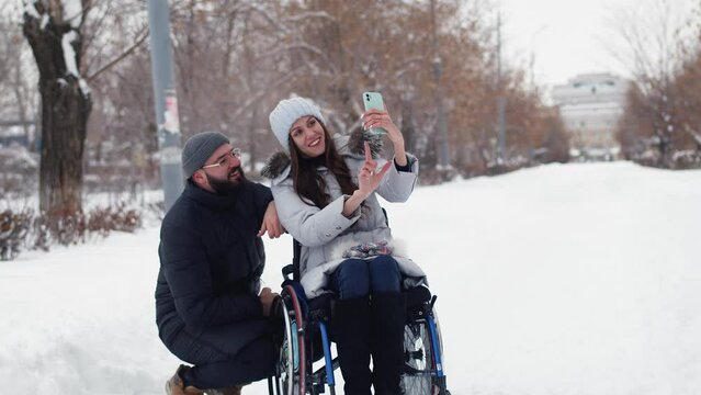 Disabled woman in wheelchairs take selfie with man on smartphone at winter park, front view. Disabled woman at wheelchairs with friend man take selfies using smartphone