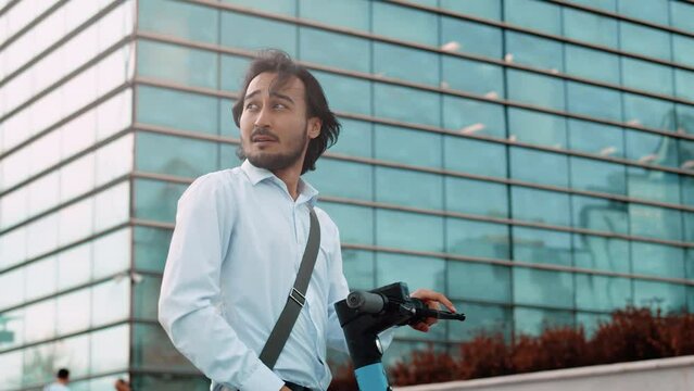 Young asian businessman in a white shirt and a shoulder bag drives up on an electric scooter to a business center and solves corporate issues on a smartphone using a speakerphone