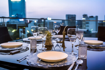 Fototapeta na wymiar A luxurious dinner is set on a marble table with glass and ceramic dishes, silverware, and a stunning cityscape view in the background. The dinner takes place on a rooftop at twilight. generative AI.