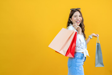 Fototapeta premium cheerful shopper smiling and holding shopping bags isolated on yellow studio background, happy young Asian pretty woman enjoy purchasing gifts, for advertising and sale promotion shopaholic concept