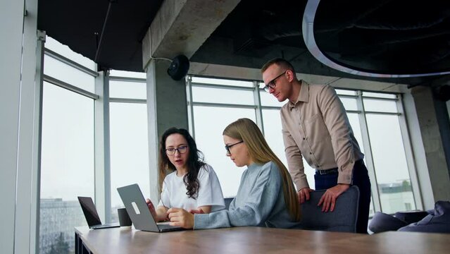 Cooperative colleagues perform internet research on the project. Male workmate stand behind females focused on the laptop.