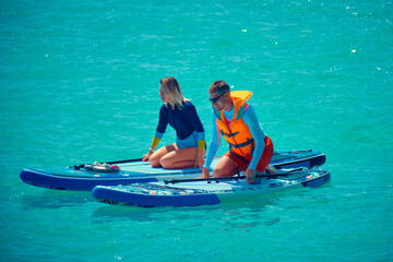 Fototapeta premium Summer outdoor seaside activities. Man and woman swim on supboards. Young man dressed in swimsuit and lifevest and young woman swimming on stand up paddle board.