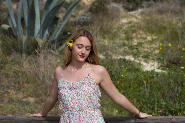 Naklejka premium Beautiful young blonde woman is wearing yellow daisy in her hair and is happy. The woman is wearing a dress with a flower pattern and is leaning on a wooden railing. Spring and nature concept.