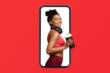 Glad young african american slim lady in sportswear and headphones with cocktail on smartphone screen