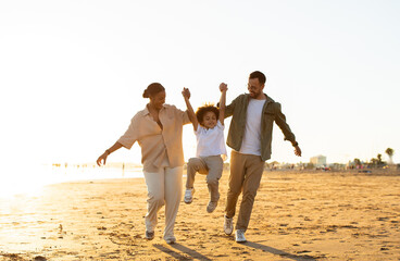 Seaside serenity concept. Happy multiracial family walking along coastline, parents lifting son up,...