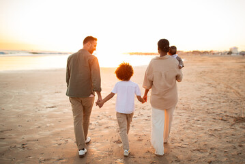 Young family taking a walk along the sea shore, holding hands with their child and walking on beach, back view