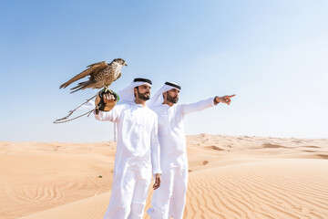 Two middle-eastern emirati men wearing arab kandura holding falcon in the desert - obrazy, fototapety, plakaty