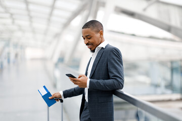 Fototapeta na wymiar Online Check-In. Smiling Black Businessman Using Smartphone While Waiting Flight At Airport
