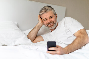 Happy grey-haired european man lying in bed with phone