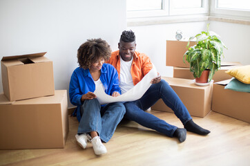 Fototapeta na wymiar Happy Black Couple Planning Home Renovation, Checking Blueprints In Their New Apartment