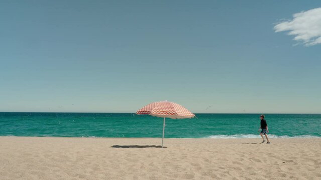 Male tourist walks along the sandy beach, against the backdrop of the sea and clear blue sky. Vibrant red parasol swinging in the wind.