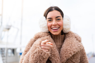 Young woman wearing winter muffs at outdoors surprised and pointing front