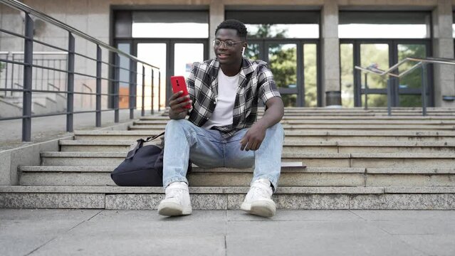 African student using mobile phone sitting on University Campus staircase - Video call