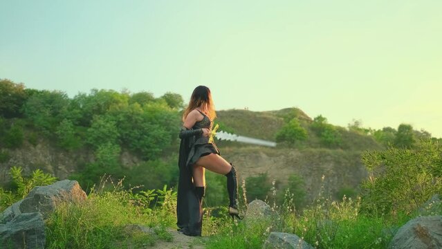 Fantasy woman warrior holds weapon in hands shouts battle cry raises sword to sky sword back rear view. Black cape costume Sexy red hair girl princess. summer nature green trees magic sun light sky