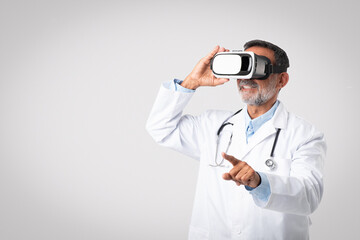 Smiling european senior doctor in white coat with stethoscope and vr glasses points finger at empty space