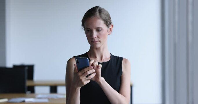 Millennial smiling businesswoman, office employee holds smartphone standing at workplace, use new professional mobile application or e-services, share messages with client in co-working. Modern tech