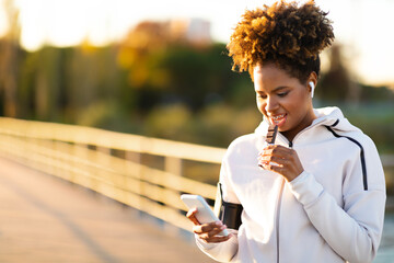 Black Woman Resting After Outdoor Training, Eating Protein Bar And Using Smartphone