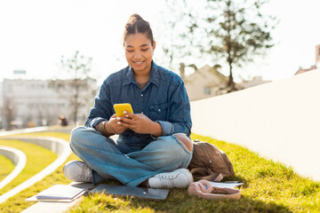 Obraz premium Happy college girl using cellphone and texting online, sitting outdoors in urban park, free space