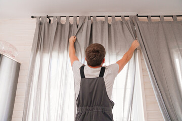 A male repairman installs a curtain rod and hangs curtains in the house.