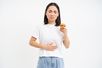 Portrait of korean woman has stomach ache, touching belly, holding cupcake, cant eat pastry, white...