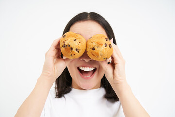 Close up portrait of funny asian girl, holds cupcakes near face, makes happy faces, white studio...