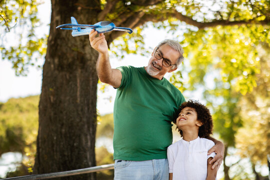 Cheerful elderly european grandfather and mixed race little boy play toy airplane in park, enjoy free time