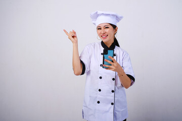 Photo of excited happy female Asian chef using mobile phone pointing on empty space or copy space for product isolated on white background