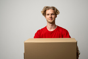 Delivery service. Young smiling courier holding cardboard box. Happy young delivery man in red t-shirt standing with parcel isolated on white background.